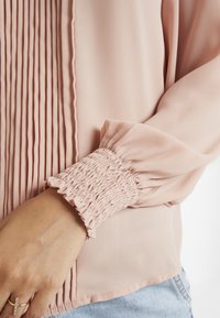 Ljusrosa blus med vertikala veck, elastiska smockärmar och slät textur. Har en rundad nederkant och subtila sömnadsdetaljer.