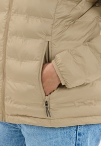 Lysebeige dunjakke med lynlås foran, quiltet design og sidelommer. Stoffet har en glat tekstur og elastiske manchetter.
