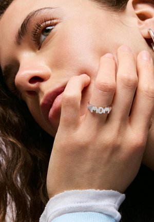 Joven mujer apoyando su rostro en la mano, lleva un anillo de plata con la palabra "MOM" en su dedo y una manga azul clara con puño.