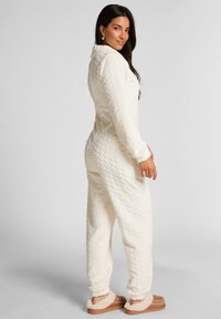 Kremowy polarowy kombinezon z teksturowanym wzorem, długimi rękawami i kołnierzykiem. W połączeniu z przytulnymi, beżowymi kapciami.