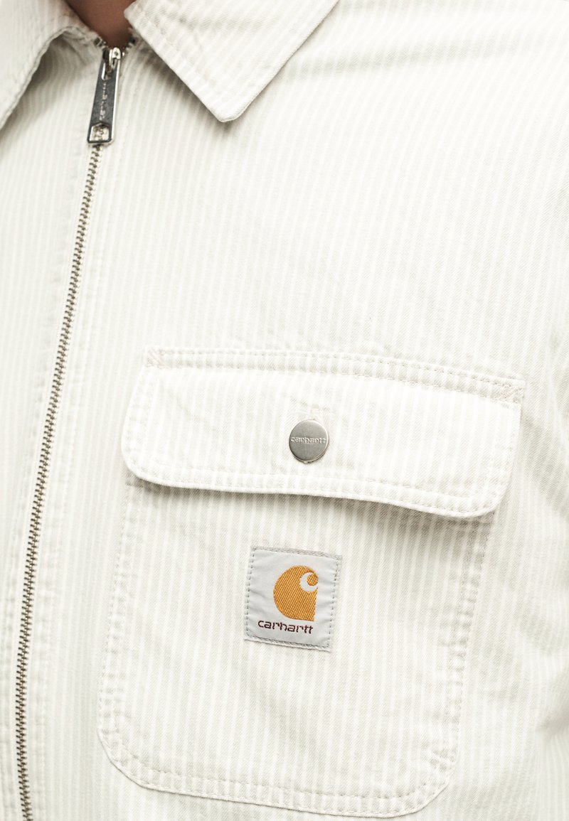 Nahaufnahme einer weiß gestreiften Jacke mit Reißverschluss, Klappentasche, die durch einen silbernen Druckknopf gesichert ist, und gelbem Carhartt-Logoaufnäher auf der Tasche.