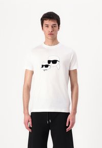 T-shirt in cotone bianco con una grafica stampata di un personaggio e un gatto con occhiali da sole neri, su uno sfondo semplice.