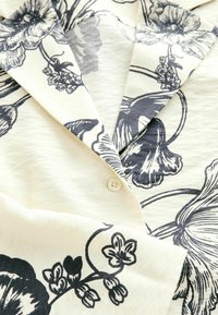 Gros plan sur un tissu de couleur crème avec un imprimé floral noir et un petit bouton blanc sur le col d'un vêtement plié.