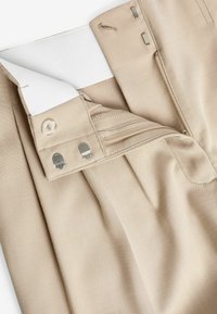 Pantalones de tela beige con un acabado texturizado. Cuentan con una cintura elástica blanca, cierre de botón y un detalle de bolsillo con cremallera.