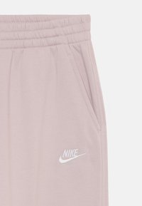 Gaiši rozā Nike sporta bikses ar elastīgu jostasvietu, sānu kabatām un baltu izšūtu logo kreisajā augšstilbā. Mīksta auduma tekstūra.