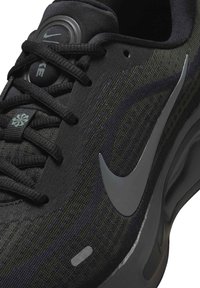 Chaussure de sport Nike noire avec une tige texturée, des accents gris, une forme arrondie et un logo proéminent avec un détail réfléchissant sur le côté.