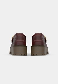 Bordeaux leren schoenen met een stevige bruine zool, voorzien van een gouden gespdetail en gestikte accenten op de achterkant.