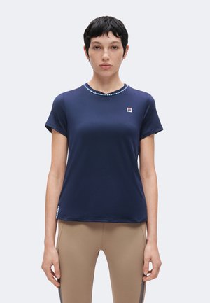 Ung person med kort sort hår iført en mørkeblå t-shirt med et lille logo på brystet og beige bukser.