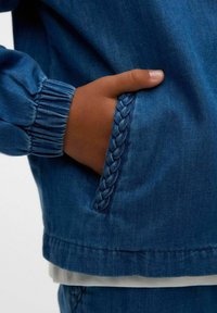 Denim jaka ar pītām akcentiem uz kabatas. Izceļas ar elastīgiem piedurknēm un gludu tekstūru vidēji zilā krāsā.