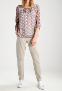 Blusa trasparente a pois in taupe con scollatura rotonda e dettaglio a chiave. Abbinata a pantaloni a sigaretta beige chiaro e sneakers bianche.