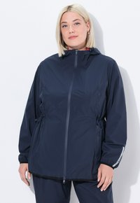 Kvinna med blont hår iklädd en marinblå huva zip-up jacka och matchande byxor, stående mot en vit bakgrund.