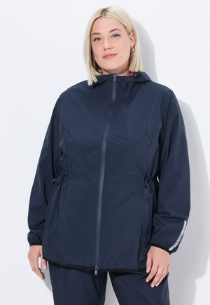 Woman with blonde hair wearing a navy blue hooded zip-up jacket and matching pants, standing against a white background.