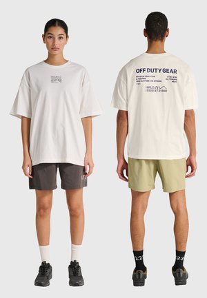 Een vrouw die naar voren kijkt en een man die naar achteren kijkt dragen beide oversized witte T-shirts met tekst, casual korte broeken, sokken en sneakers tegen een effen achtergrond.