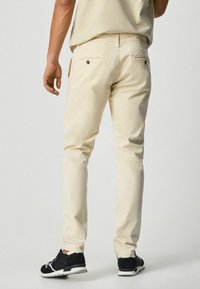 Beige slim-fit byxor med bakfickor och bruna knappar, i kombination med svarta sneakers. Tyget ser jämnt ut med en minimalistisk design.