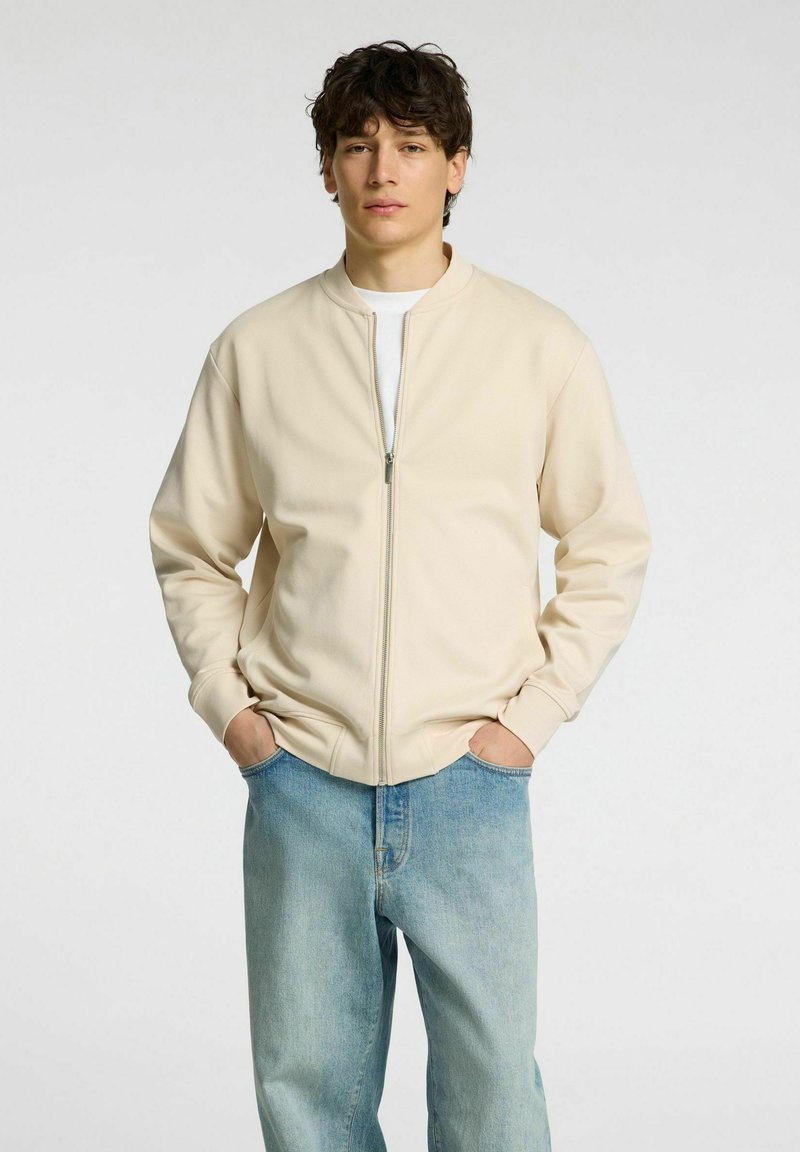 Beige színű zipzáros dzseki kerek nyakkivágással, bordázott mandzsettákkal és laza szabással, világoskék farmernadrággal párosítva. Simas texture.
