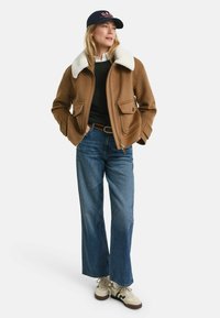 Braune Wollbomberjacke mit weißem Kunstfellkragen, schwarzer Strickpullover darunter, blaue weit geschnittene Jeans und beigefarbene Sneaker. Dunkelblaue Mütze mit Logo.