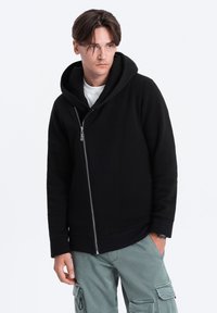 Zwarte ritsshirt hoodie met een grote capuchon, textuurstof en schuine rits. Gecombineerd met lichtgroene cargobroek die een moderne pasvorm toont.