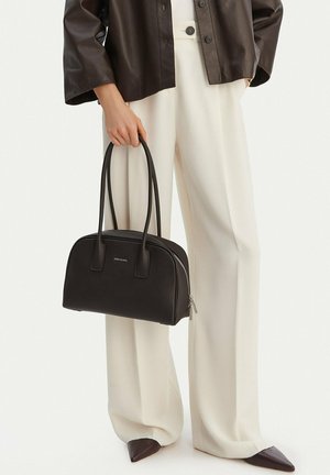 Žena v širokých bílých kalhotách a tmavé bundě, držící černou koženou kabelku s dlouhými uchy a zipovým zavíráním.