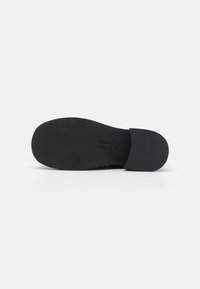 Sole di una scarpa nera senza lacci con una superficie liscia e opaca. Presenta una forma arrotondata e un design minimalista, con un logo del marchio impresso in modo discreto.