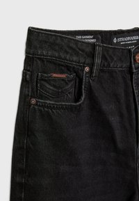 Jeans de mezclilla negra con un diseño clásico de cinco bolsillos, remaches de cobre y una etiqueta tejida en el bolsillo trasero. Textura suave y tiro medio.