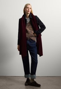 Jersey marrón de punto debajo de un abrigo oscuro, combinado con unos jeans de mezclilla doblados y zapatos oscuros. Los accesorios incluyen una larga bufanda burdeos.