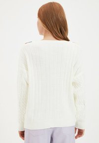 Femme aux longs cheveux roux portant un pull blanc côtelé et tricoté en torsades, debout de dos, assorti à un pantalon violet clair.