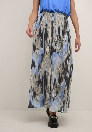 Frau trägt einen fließenden, knöchellangen Rock mit abstrakten Mustern in Blau, Schwarz und Beige, dazu schwarze klobige Loafers und ein blaues Oberteil.