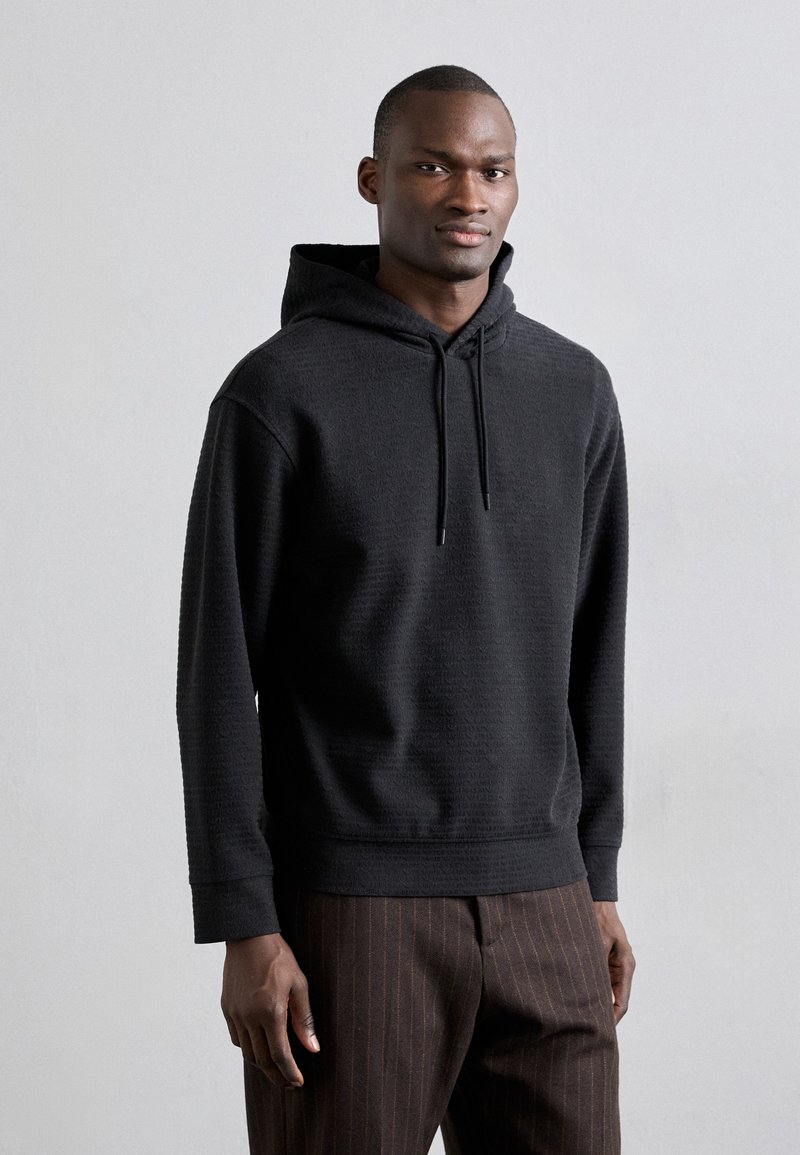 Svart huvtröja med ett texturerat mönster, med dragsnöre och ribbade muddar, i kombination med bruna ränderbyxor.