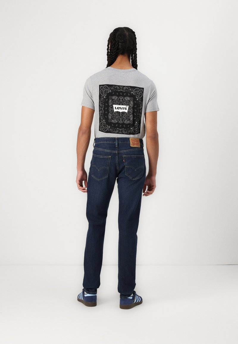 Camiseta gris con diseño de paisley negro y logo de "Levi's", combinada con jeans azul oscuro y zapatillas a rayas azules.