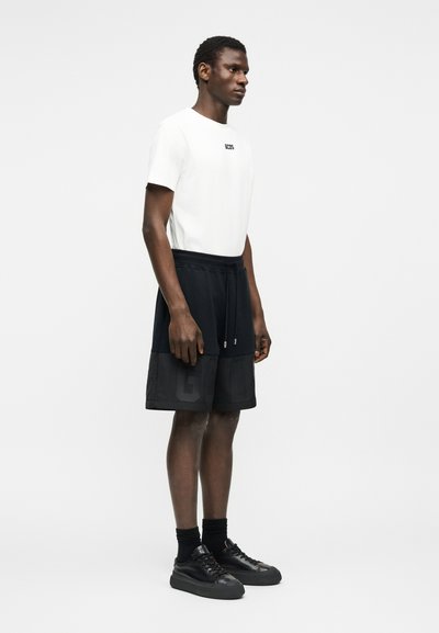 Άνδρας που φοράει λευκό κοντομάνικο μπλουζάκι με λογότυπο, μαύρο σορτς με κορδόνι, μαύρες κάλτσες και μαύρα αθλητικά παπούτσια, στέκεται πλαγίως σε απλό φόντο.