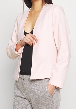 Kvinde iført en lyserød blazer over en sort top og grå teksturerede bukser med den ene hånd i lommen og den anden justerende blazeren.