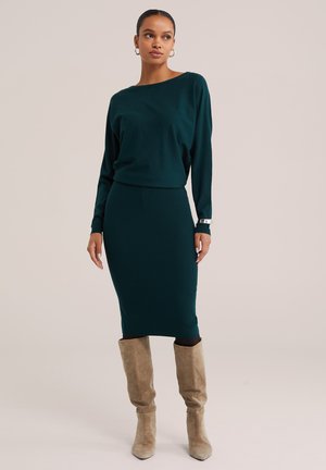 Femme portant un haut à manches longues vert foncé et une jupe ajustée assortie avec des bottes en daim beige montant jusqu'aux genoux sur un fond neutre.