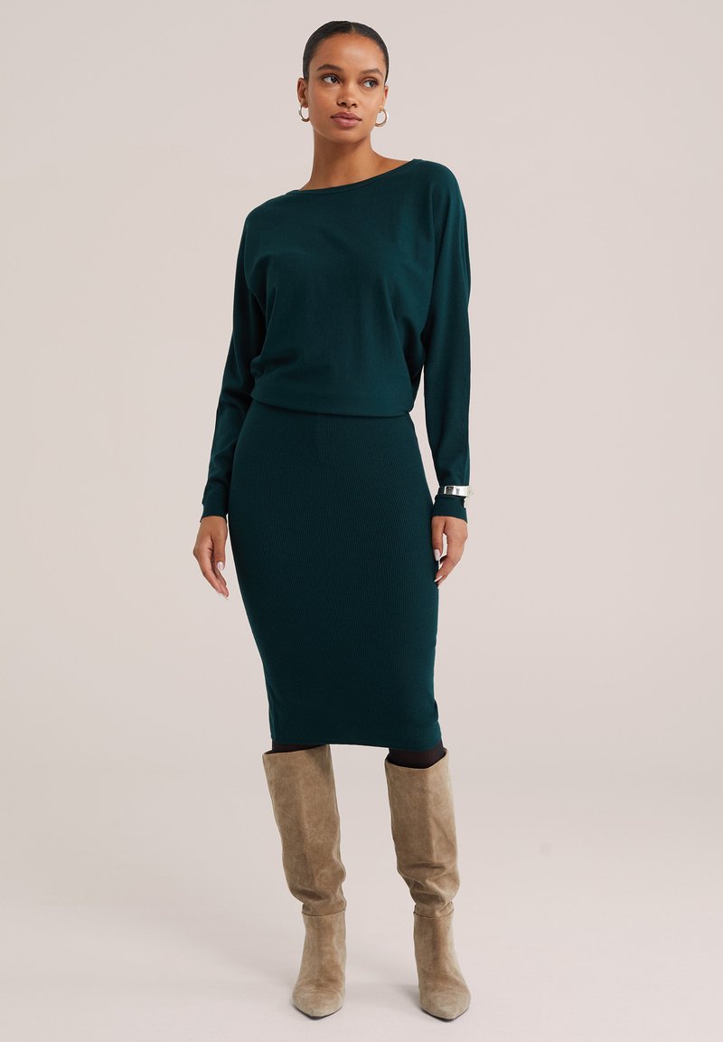 Femme portant un haut à manches longues vert foncé et une jupe ajustée assortie avec des bottes en daim beige montant jusqu'aux genoux sur un fond neutre.