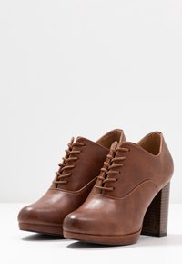 Scarpe in pelle marrone con tacco, caratterizzate da una punta arrotondata, chiusura con lacci e un tacco a blocco strutturato. Finitura liscia con dettagli di cucitura discreti.