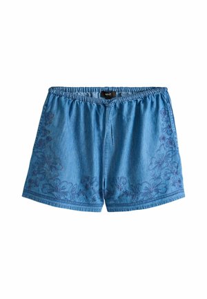 Nahaufnahme einer Person, die ein blaues Denim-Outfit mit Blumenstickerei trägt und die Kordel an einer hochtaillierten Hose bindet.