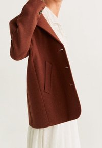 Manteau en mélange de laine marron avec un large col, une fermeture à un bouton, une poche latérale et une silhouette structurée, porté par-dessus une robe blanche.