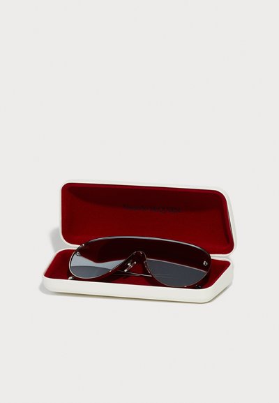 Sunglasses in a red-lined white case; features a curved, visor-like dark lens design with metal accents and a prominent frame shape.