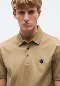 Béžové polo tričko s golierom a tromi gombíkmi, s čiernou logo nášivkou na hrudi. Hladká bavlnená látka s krátkymi rukávmi.