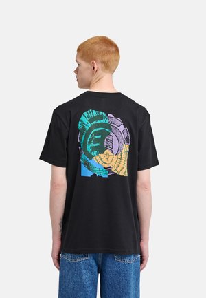 Giovane con capelli rossi corti che indossa una t-shirt nera con un disegno circolare astratto multicolore sulla schiena e jeans blu.