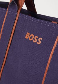 Borsa a spalla viola in tela con manici a righe marroni e logo "BOSS" in rilievo di una tonalità più chiara. Texture liscia, design robusto.