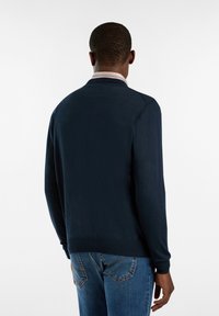 Marineblauer Pullover mit einer glatten, weichen Textur, ausgestattet mit einem gestreiften Kragen und langen Ärmeln, getragen über blauen Jeans.