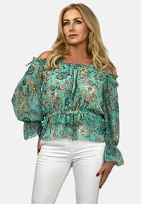 Turkusowa bluzka typu off-the-shoulder z falbaniastymi rękawami, w wzór paisley, z elastyczną talią. W zestawieniu z białymi dopasowanymi spodniami.