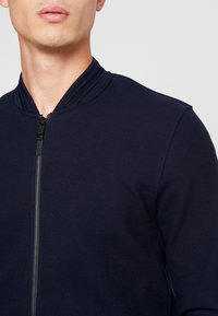 En man som bär en marinblå strukturerad zip-up jacka med ribbad krage mot en enfärgad ljusgrå bakgrund.