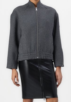 Personne portant une veste zippée gris foncé avec taille élastique et une jupe en cuir noire zippée sur un fond blanc uni.