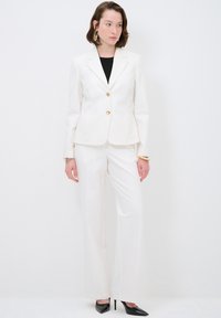 Conjunto de traje blanco que incluye un blazer ajustado con botones dorados y pantalones de pierna ancha. Una blusa negra y tacones negros puntiagudos completan el look.