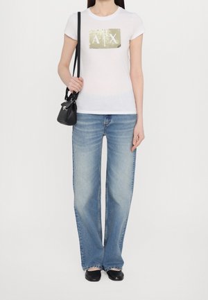 Hvid T-shirt med metallisk guld "A|X" logo, kombineret med lyseblå vide jeans og sorte flade sko, der holder en lille sort taske.