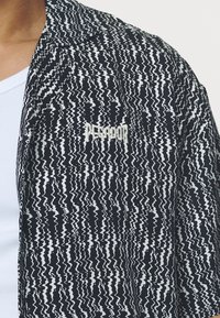 Camisa preta e branca com padrão, apresentando um design ondulado, feita de tecido leve, com um logo bordado na área do peito esquerdo.