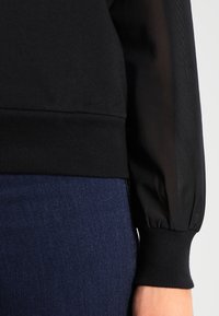 Felpa nera con maniche a sbuffo trasparente, polsini e fascia in vita a coste. Abbinata a denim blu scuro, che mette in risalto texture a contrasto.