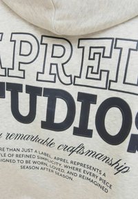 Světle šedá mikina s kapucí se silným černým textem "APREL STUDIOS" a menším dekorativním textem. Měkká tkanina s hladkým povrchem.