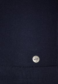 Navy gebreide trui met een gestructureerde stof, voorzien van een geribbelde zoom en een ronde metalen logo-accent aan de onderkant rechts.